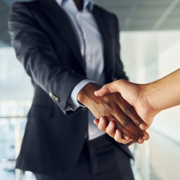 Doing handshake with customer. Young african american businessman in black suit is indoors.